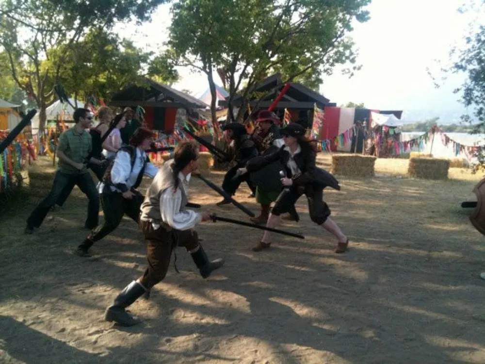 Players in period costume fighting with foam swords in a sunlit dirt courtyard ringed with colorful banners and hay bales
