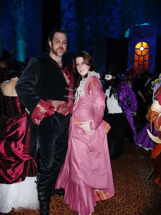 A costumed couple at a masquerade ball, a man in a black coat with a red vest and a woman in a rose-pink gown with a lace collar