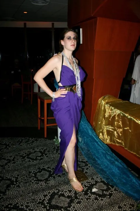 A woman in a purple draped gown with a long teal train, layered pearl necklaces, and gold sandals