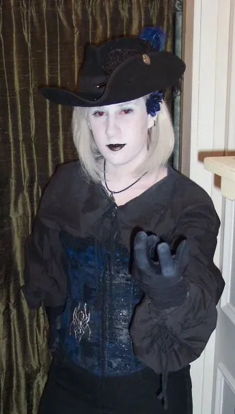 A player with white face paint, black lips, and a black feathered hat, in a corset and black shirt with a gloved hand raised